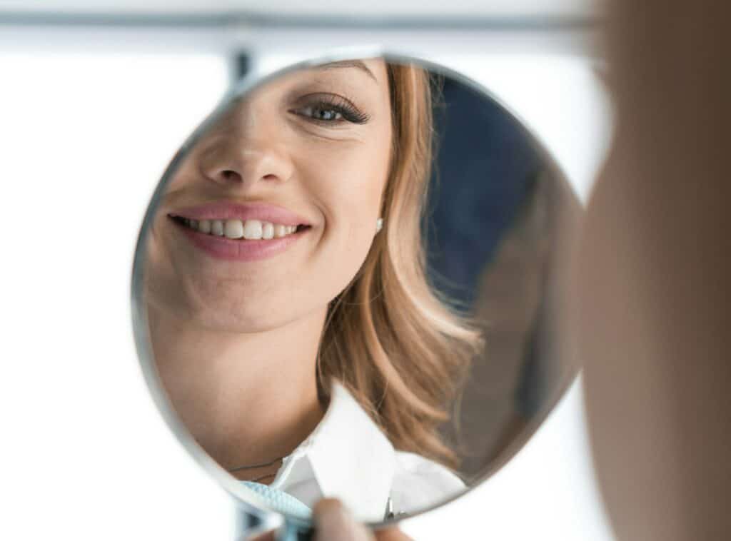 In mirror can see distinct improvements in condition teeth after visiting dentist. Woman is glad